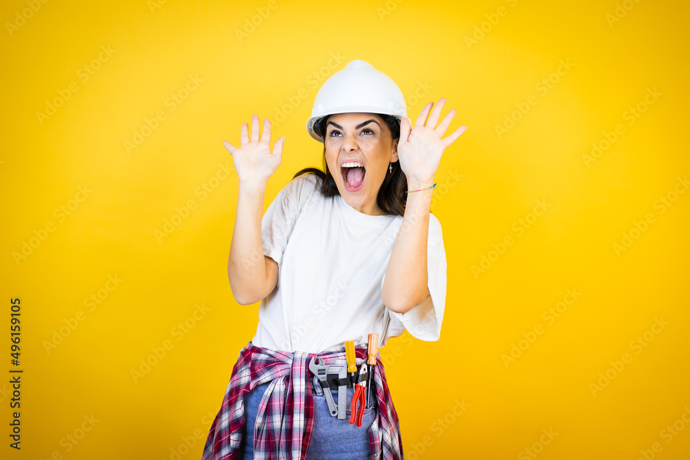 Young caucasian woman wearing hardhat and builder clothes over isolated yellow background scared with her arms up like something falling from above