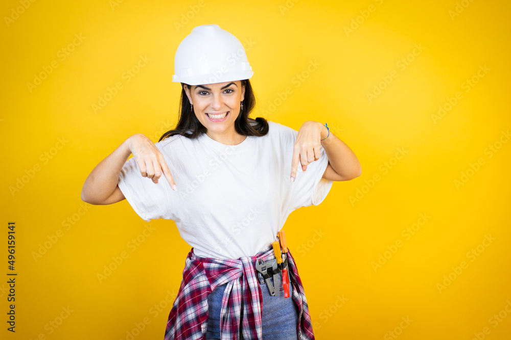 Young caucasian woman wearing hardhat and builder clothes over isolated yellow background surprised, looking down and pointing down with fingers and raised arms