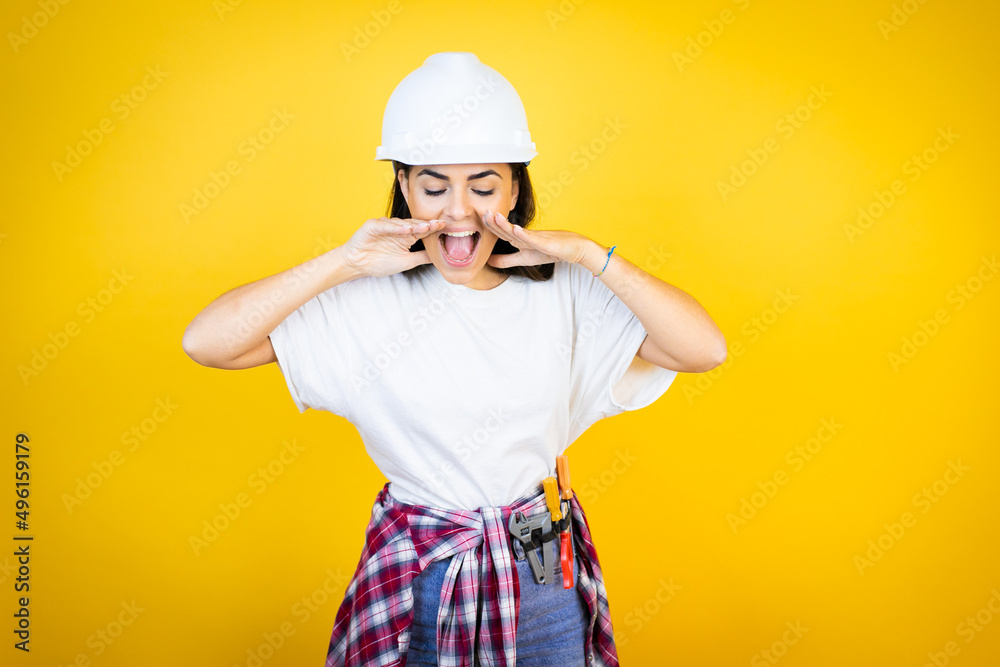 Young caucasian woman wearing hardhat and builder clothes over isolated yellow background shouting and screaming loud down with hands on mouth