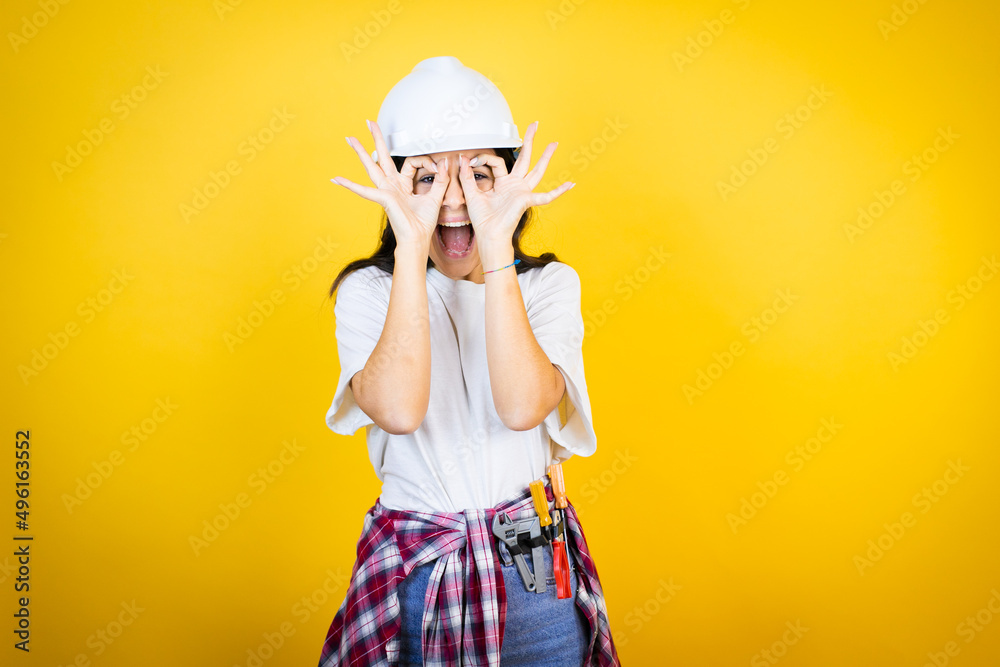 Young caucasian woman wearing hardhat and builder clothes over isolated yellow background doing ok gesture shocked with smiling face, eye looking through fingers