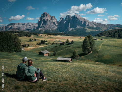Couple sitting on meadows with wooden cabins at Alpe di Siusi during summer with view to mountains of Plattkofel and Langkofel in the Dolomite Alps in South Tyrol, Italy.