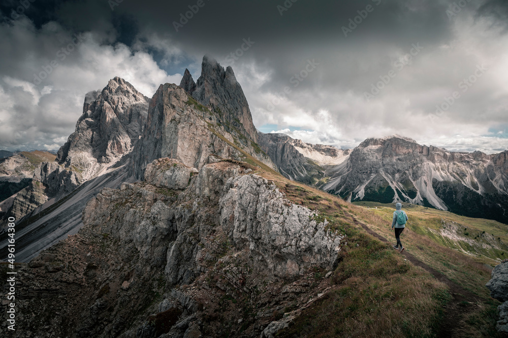 Fototapeta premium Girl hiking along cliffs of dramatic mountain peaks of Seceda with clouds in the European Dolomite Alps, meadow in the foreground, steep cliff, South Tyrol Italy.