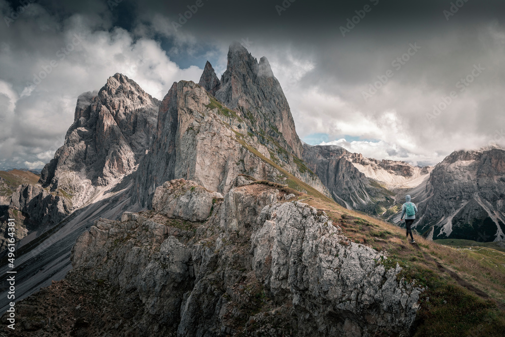 Girl hiking along cliffs of dramatic mountain peaks of Seceda with clouds in the European Dolomite Alps, meadow in the foreground, steep cliff, South Tyrol Italy.