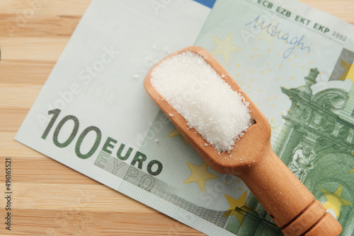 White sugar in a wooden spoon. One hundred euro. Background light tree. Deficient item. Harm and benefit, high-calorie product. Copyspace.