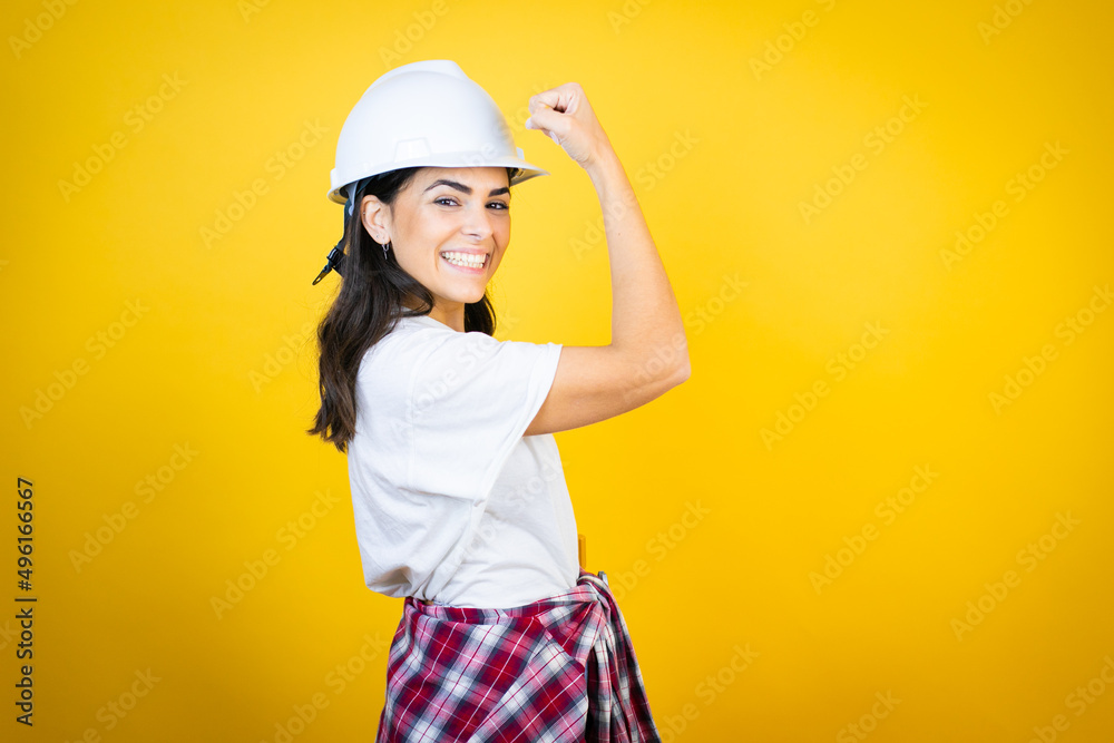 Young caucasian woman wearing hardhat and builder clothes over isolated yellow background feeling happy, satisfied and powerful, flexing fit and muscular biceps, looking strong after the gym