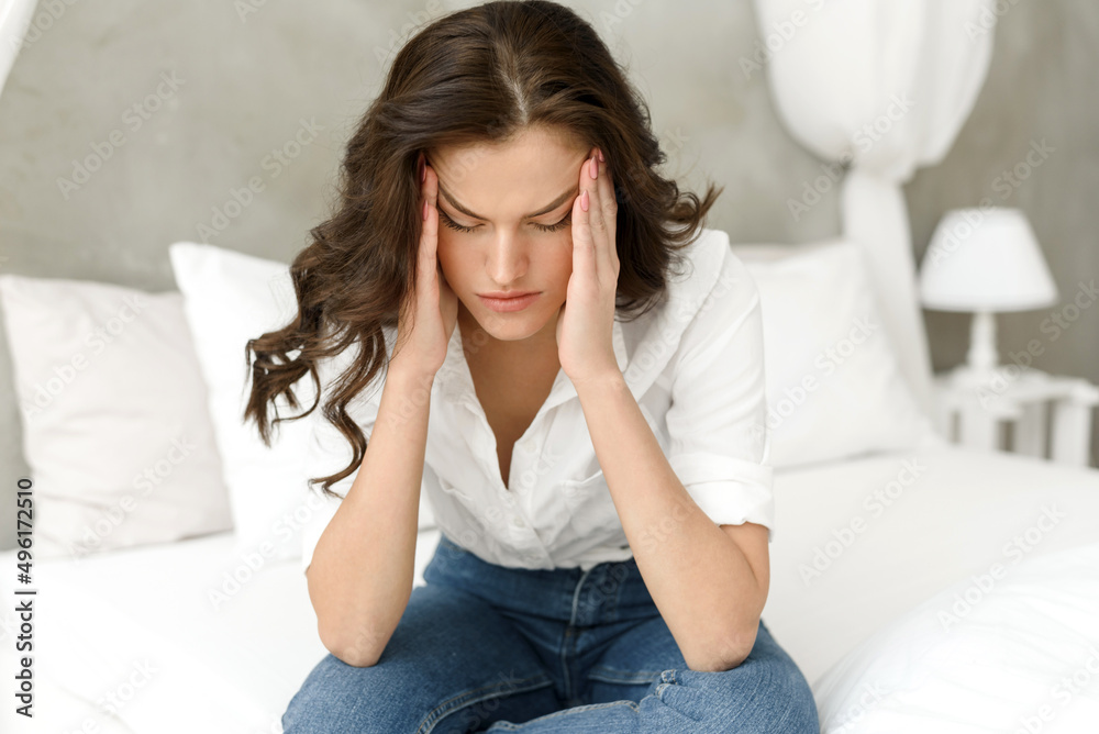 Unhappy woman touching her head, sitting on the bed at home, having a headache. Young frustrated woman thinks about problems, feels lonely and sad Unhappy woman touching her head, sitting on the bed at home, having a headache. Young frustrated woman thinks about problems, feels lonely and sad
