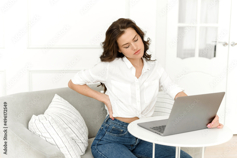 Tired young woman freelancer sitting at the table, touching her back, suffering from a spasm of the lower spine, severe back pain, incorrect posture concept Tired young woman freelancer sitting at the table, touching her back, suffering from a spasm of the lower spine, severe back pain, incorrect posture concept