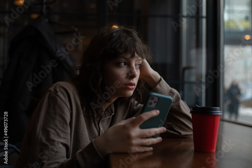 A girl with curly hair uses a smartphone while sitting in a cafe by the window.