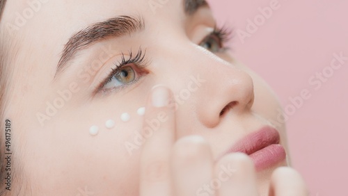 Extreme close-up of young beautiful fit Caucasian skin care woman applies cream under her eye on pink background | Eye cream commercial concept