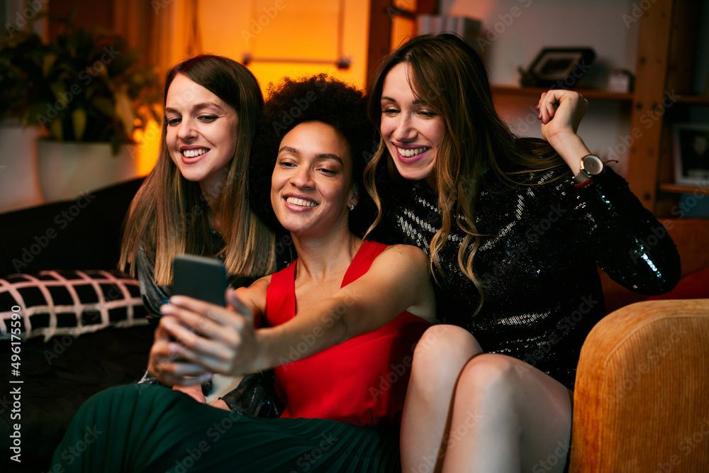 Three multicultural female friends sit in the living room in the evening and take selfies at home party.