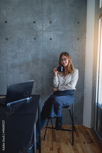Fotografie A pretty casual businesswoman with glasses sits on the chair between the desk and window in the corner of the chancellery