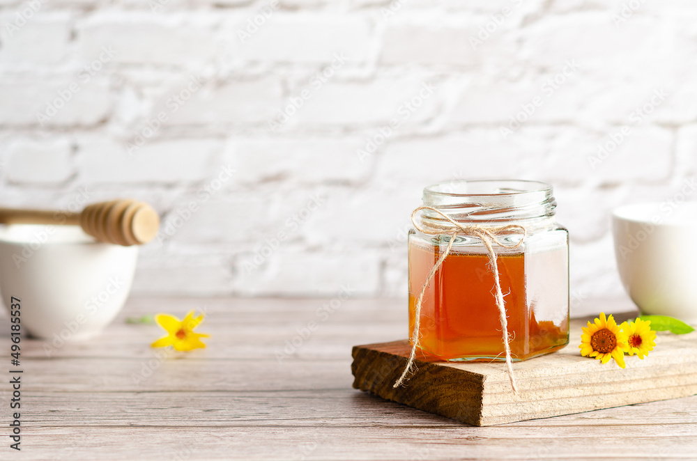 Honey in a jar on a piece of wood, a honey dripper, white bowls and some marigolds, on a wooden table and a white wall. Honey in a jar on a piece of wood, a honey dripper, white bowls and some marigolds, on a wooden table and a white wall.