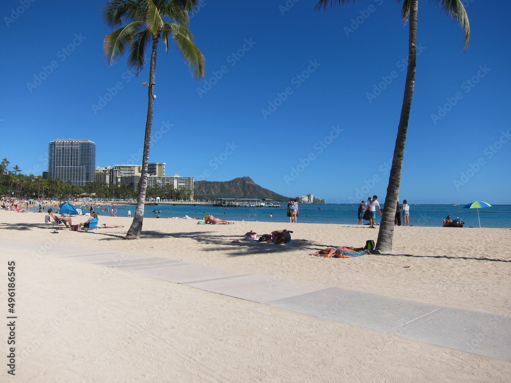 View of Kahanamoku Beach, Waikiki. View of the horizon with palm trees, Diamond Head mountain summit, clear blue sky, the skyline, and a walkaway across the beige sand. Oahu, Hawaii.