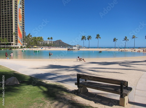 View of Kahanamoku Beach, Waikiki, from behind a wooden bench. View of the horizon with palm trees, Diamond Head mountain summit, clear blue sky, green grass, buildings and the lake.