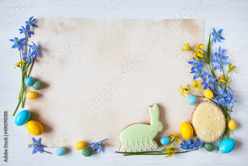 Easter greeting card. White wooden background, blue and yellow spring flowers, egg shaped gingerbread cookies, bunny, candies and paper for text.