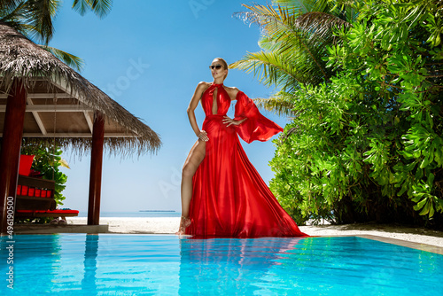 Luxury fashion. Elegant fashion model. Stylish female model in red long gown dress on the Maldives beach. Elegance. Classy woman in amazing red dress near the pool. Couture. Vogue.