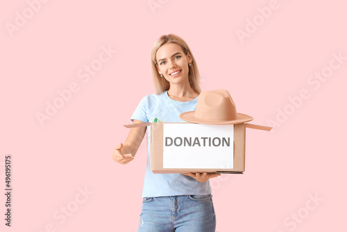 Young woman holding box with donation clothes on pink background