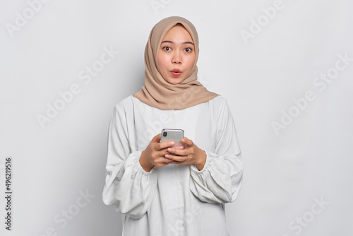 Amazed young Asian Muslim woman using a mobile phone and looking at camera isolated over white background