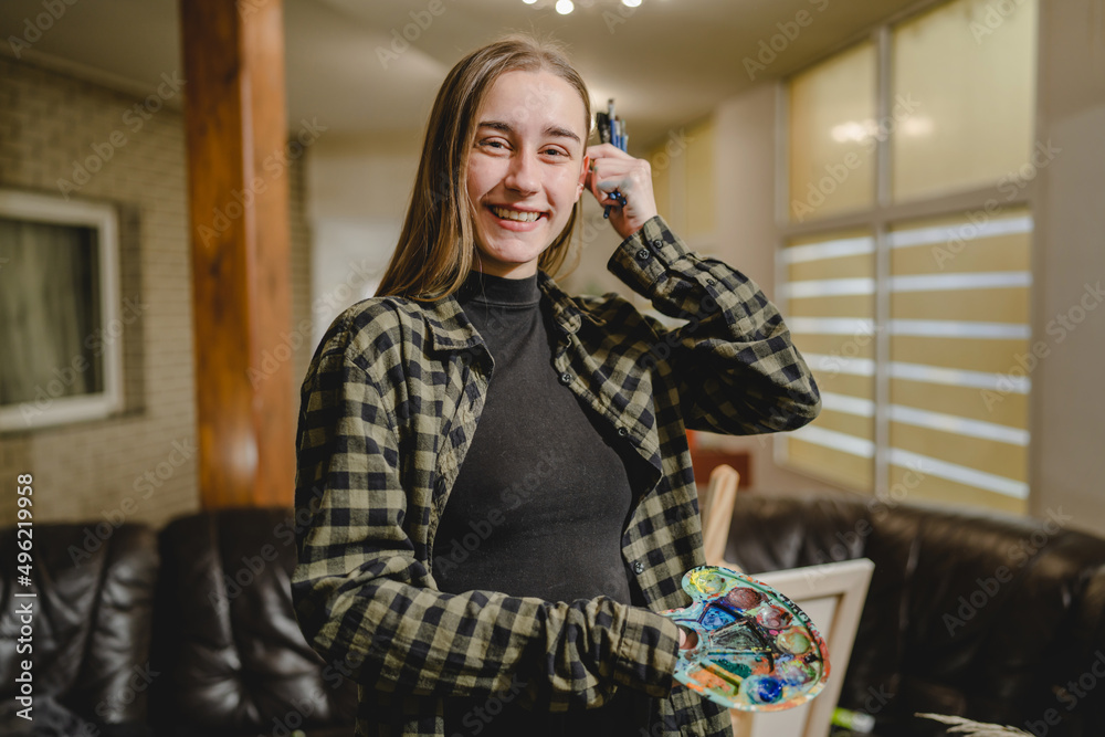 Fototapeta premium One young caucasian woman female artist professional or amateur painter student standing at home with color palette and paintbrushes in hands posing with copy space waist up