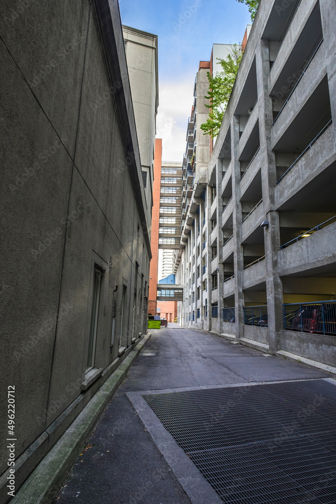 A deserted lane way with a parking garage on the right side. Garbage bins and an elevated walkway are in the distance.