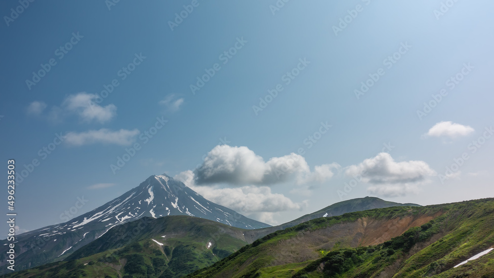 A beautiful conical volcano on a blue sky background. Snow lies on the slopes. Clouds over the top. Green hills in the foreground. Kamchatka. Stratovolcano Vilyuchinsky