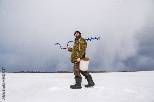 A real fisherman walks on the ice of the lake with a box and a drill.
