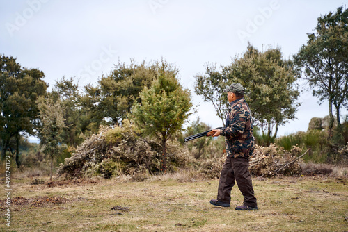 Male hunter in camouflage outerwear and cap carrying a gun walking in the field