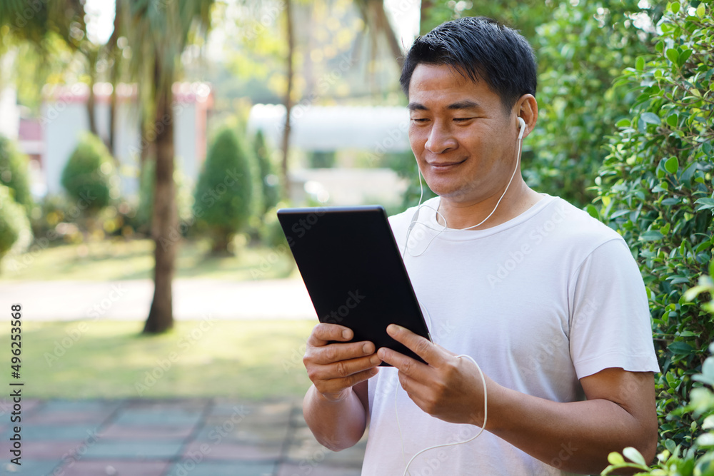 Fototapeta premium Asian man listens and watch video clips on smart tablet at the park. Concept : Give time for yourself to relax with wireless technology. Internet of things. Social media. 
