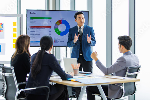 Asian professional successful businessman in formal suit standing showing presenting explaining report investment graph chart data from computer monitor in meeting room to businesswoman colleagues photo