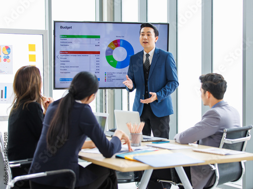 Asian professional successful businessman in formal suit standing showing presenting explaining report investment graph chart data from computer monitor in meeting room to businesswoman colleagues