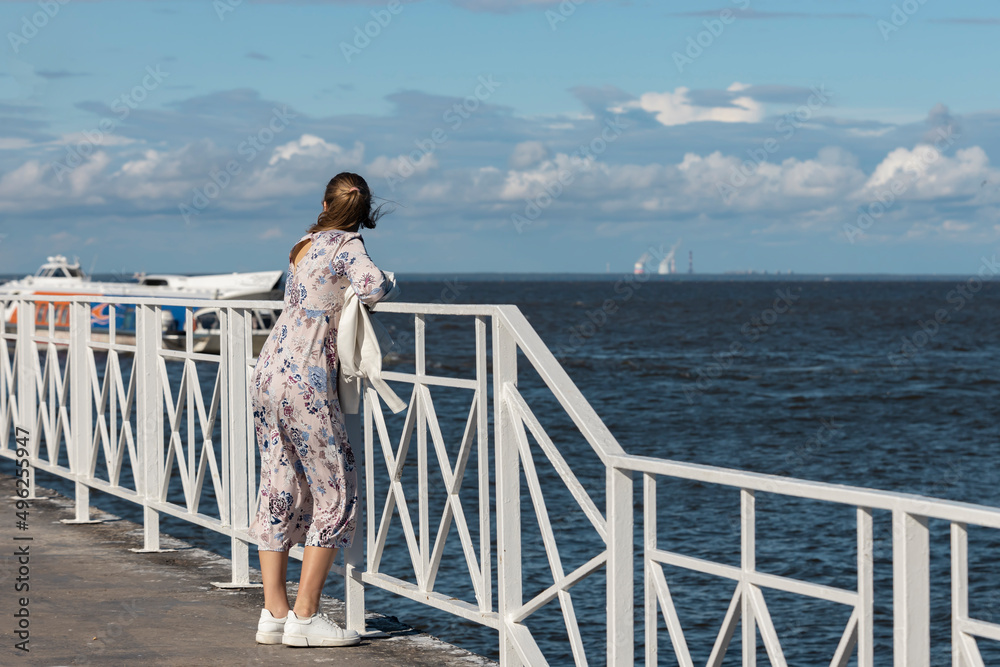 Young girl stands on a sea pier on a sunny summer day and looks into the distance.
