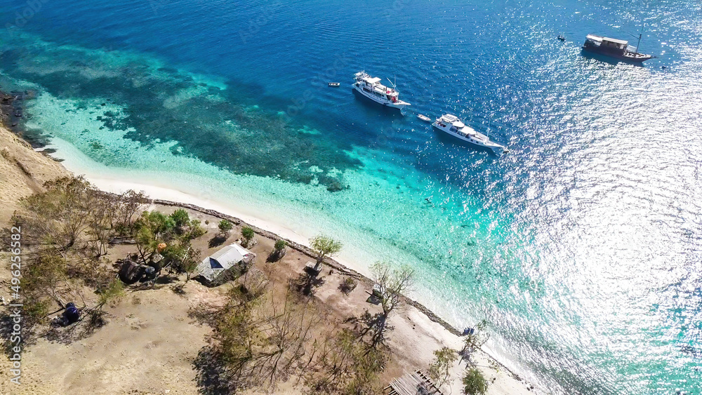 Top down drone shot of a paradise island with some boats anchored around in Komodo National Park, Flores, Indonesia. Brownish island turns into white sand beach and further into turquoise and navy sea