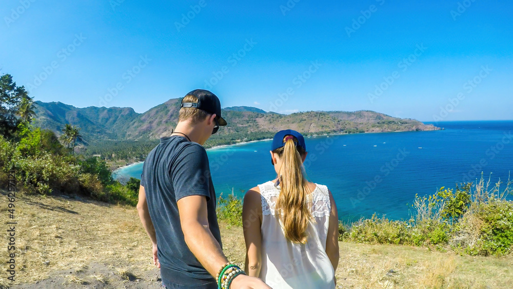 A couple walking towards an edge of a cliff. ON the bottom there is an idyllic white sand beach on Lombok, Indonesia. The beach line is overgrown with trees. Calmness and solitude while travelling.