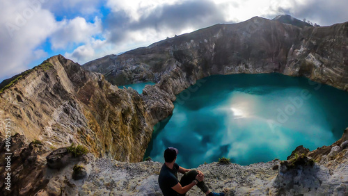 Man sitting at the volcano rim and watching the Kelimutu volcanic crater lakes in Moni, Flores, Indonesia. Man is relaxed and calm, enjoying the view on lake shining with many shades of turquoise