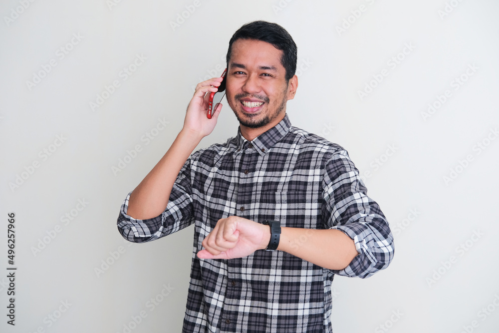 Adult Asian man smiling to the camera while make a phone call and showing his watch Adult Asian man smiling to the camera while make a phone call and showing his watch