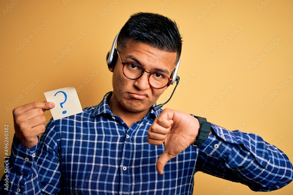 Young latin call center agent man holding question mark reminder paper using headset with angry face, negative sign showing dislike with thumbs down, rejection concept Young latin call center agent man holding question mark reminder paper using headset with angry face, negative sign showing dislike with thumbs down, rejection concept