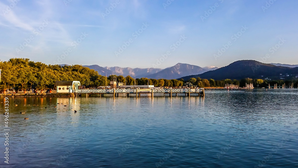 A pier on the lake captured during golden hour. In the back there is a lot of mountain ranges. Trees are slowly turning golden. The surface of the lake is very calm.