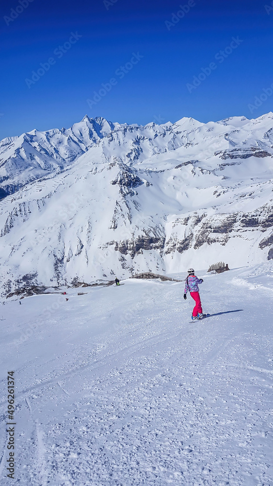 A snowboarder going down the slope in Heiligenblut, Austria. Perfectly groomed slopes. High mountains surrounding the man wearing yellow trousers and blue jacket. Girl wears helm for the protection.