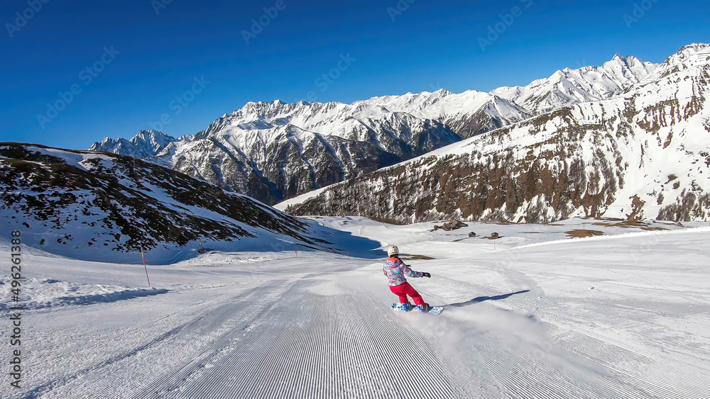 A snowboarder going down the slope in Heiligenblut, Austria. Perfectly groomed slopes. High mountains surrounding the man wearing yellow trousers and blue jacket. Girl wears helm for the protection.