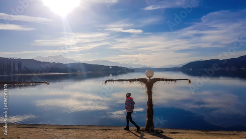 A girl in a winter jacket walks along the lakeside. There is a scarecrow next to the shore. The surface of the lake is still and calm. There are tall Alps chains in the back. Sunny cold day.