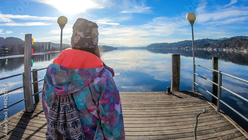 A girl wearing a beanie walks along the pier on a lake and admires the view. Calm surface of the lake reflects the sky. Girl is enjoying her time and beautiful weather. Happy wintery moment. photo