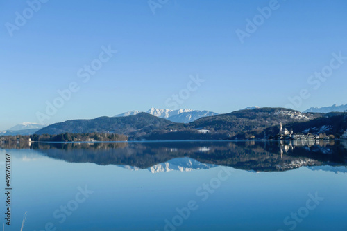 A view on a lake and Alps in the back. The calm surface of the lake is reflecting the mountains, sunbeams and clouds. Little city located at the lake side. Church with bell tower in the middle