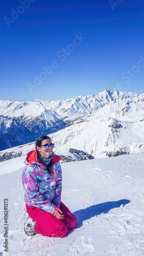 A snowboarder girl sitting on a slope admiring the tall Alps in front of her. Slopes are perfectly graveled. Perfect weather for a ride. High alpine snowboarding. Girl wears a helmet for protection.