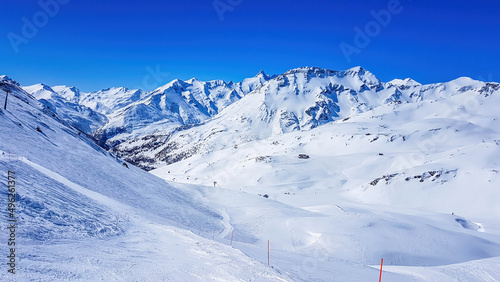 A beautiful and serene landscape of mountains covered with snow. Thick snow covers the slopes. Clear weather. Perfectly groomed slopes. Massive ski resort. Red ski poles on the side of the trail.