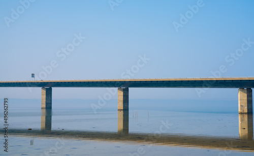 Long highway bridge spanning over a bay, with traffic driving on it. Calm water and morning haze.