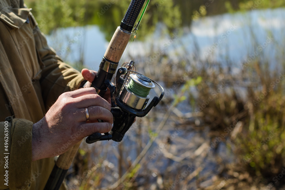 In the season of spring, a man fisherman in a fabric windbreaker holds a fishing rod, spins the reel, pulling up the fishing line, pulls out the caught fish, space for text. In the season of spring, a man fisherman in a fabric windbreaker holds a fishing rod, spins the reel, pulling up the fishing line, pulls out the caught fish, space for text.