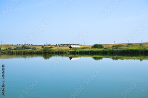 Sunny day landscape of a boundless blue lake with a lonely house for rest and unity with nature among green sedge against a cloudless sky, copy space.