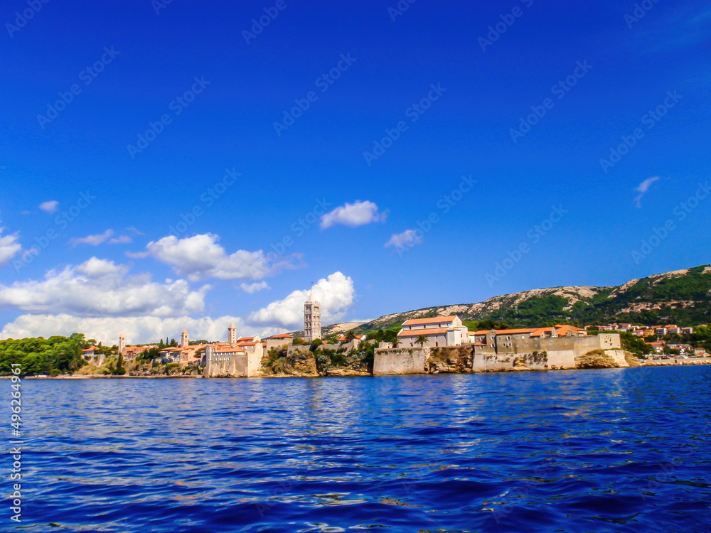 Sea view on Croatian city, Cres. The city is build directly by the sea. Thick walls emerge high up from the sea line, defence building. There is a church tower, towering above other constructions.