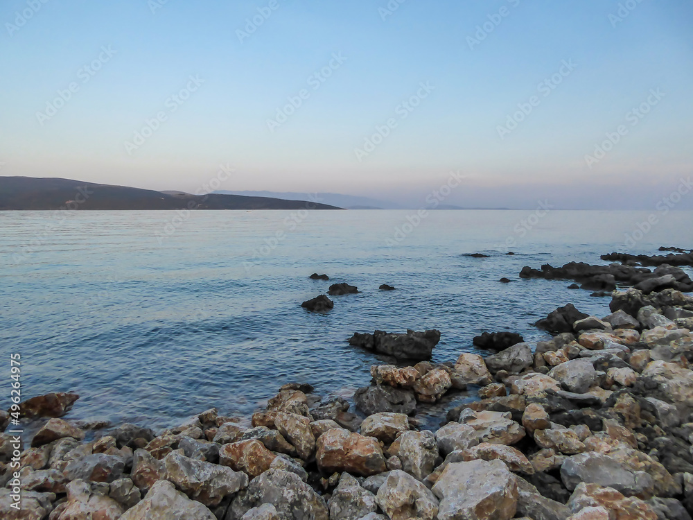 Stony beach in Krk, Croatia. The Mediterranean Sea has a turquoise color. Surface of the sea is calm, there are no waves. In the back there are some mountains.  Soft colors of the sunset.