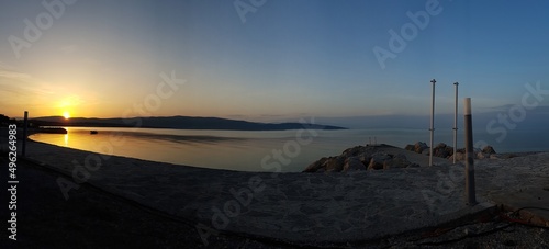 A beautiful sunrise over the beach in Krk, Croatia. Sun is rising from behind the hill, turning sky orange. Calm surface of the sea reflects the sunbeams. Swimming facilities to the right side. © Chris